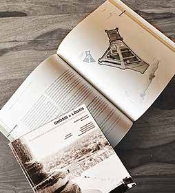 The printed master’s thesis “Gnòsis e Lògos” lying open on a wooden table: a double-page spread with an axonometric view of the fortress, below it the cover with a historical photograph
