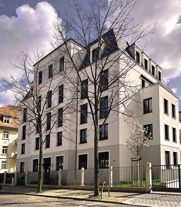 Photo of the completed Therese apartment villa: white corner façade with mansard roof behind bare street trees and a fence