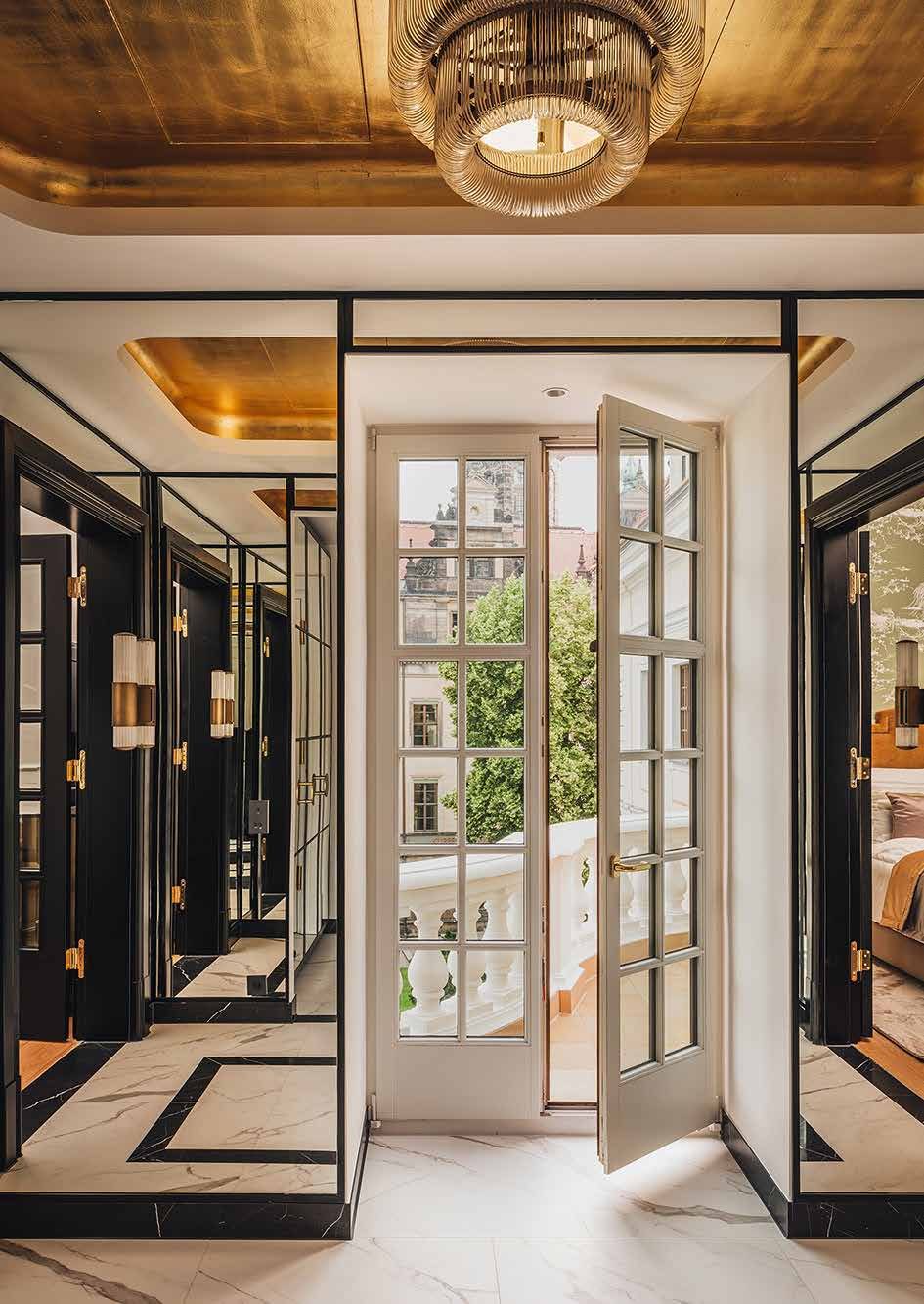 A room in the refurbished Taschenbergpalais: gilded ceiling with a glass chandelier, black-framed mirrored and glazed doors, marble floor with black inlays and an open white French door onto the balcony, overlooking Dresden’s old town