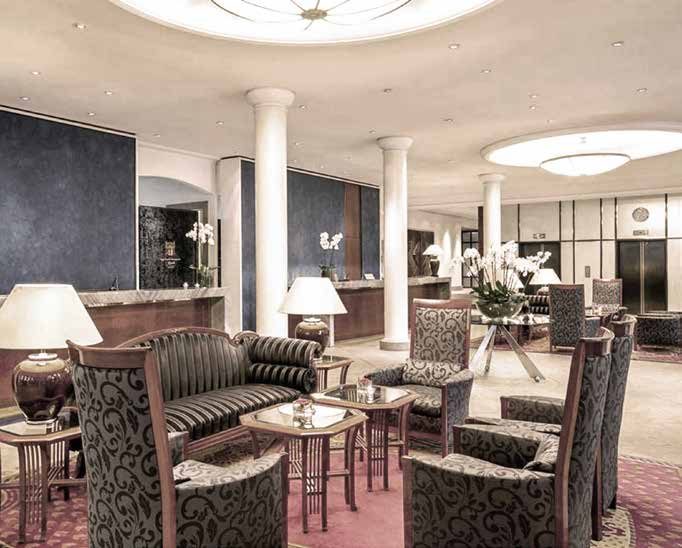 The lobby before the refurbishment: a seating group with a striped sofa and ornately patterned upholstered armchairs on a red carpet, table lamps, white columns and, behind, the reception desk against dark blue walls