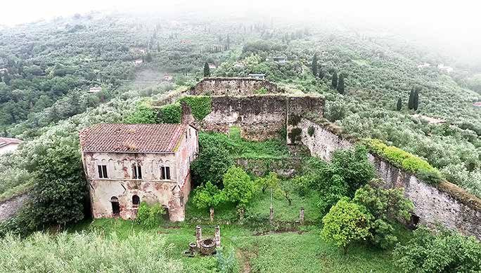 Aerial view of the existing site: the dilapidated Palazzo Guinigi with its red tiled roof inside the overgrown fortress of Pietrasanta, wooded hills behind