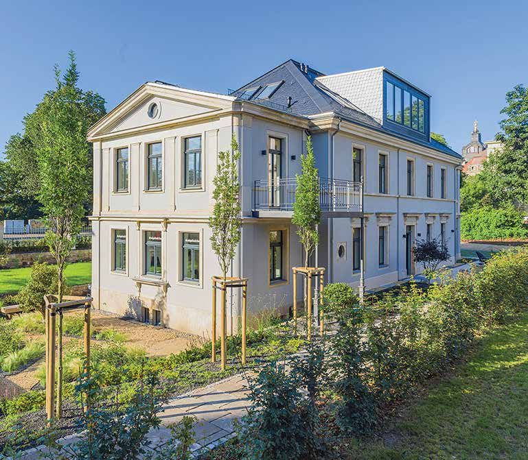 Nachher: die sanierte Villa von der Gartenseite, hell verputzt, mit neuem Schieferdach, verglastem Dachaufbau, Balkon und frisch gepflanzten Bäumen