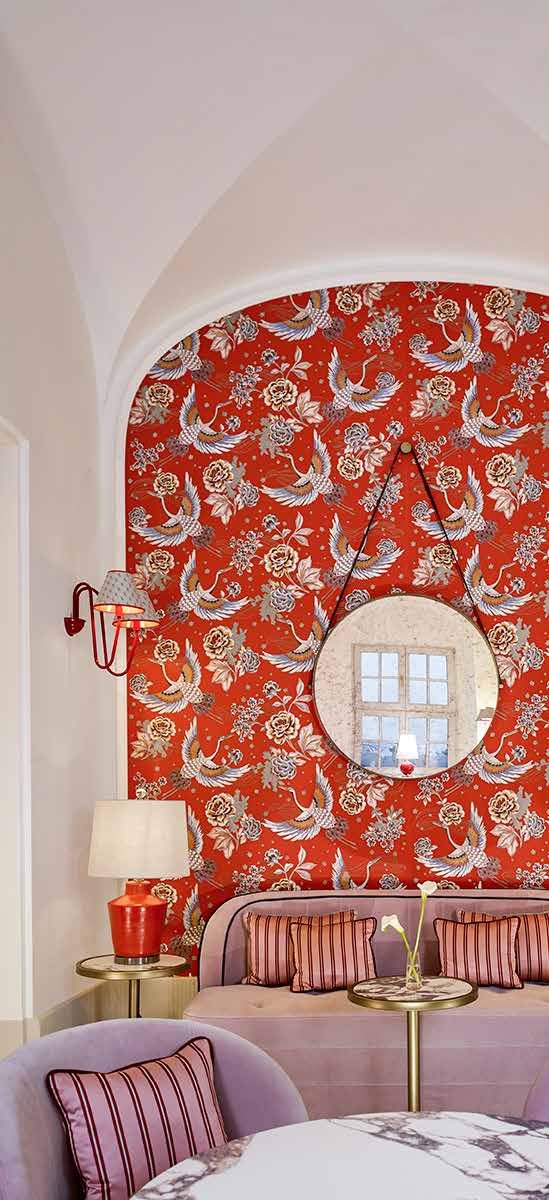 Seating corner in the patisserie: red wallpaper with a bird motif, a round mirror, a pink sofa with striped cushions and a table lamp