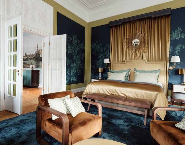 Bedroom of the Royal Suite: a bed in front of a gold curtain with a sunburst mirror, dark blue wall panels with botanical motifs in gilt frames, cognac-coloured upholstered armchairs on a blue carpet; through the open double door, a glimpse of the Dresden view in the adjoining room